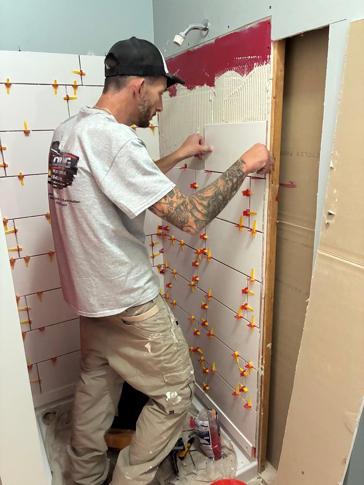 Tattooed worker in black cap installing tile on bathroom wall with orange spacers and red grout lines visible