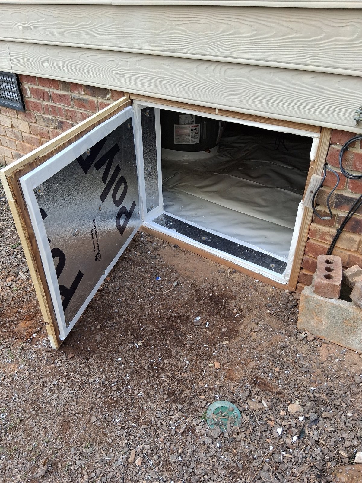 Open basement window well with white frame attached to red brick foundation, set into gravel ground against white siding
