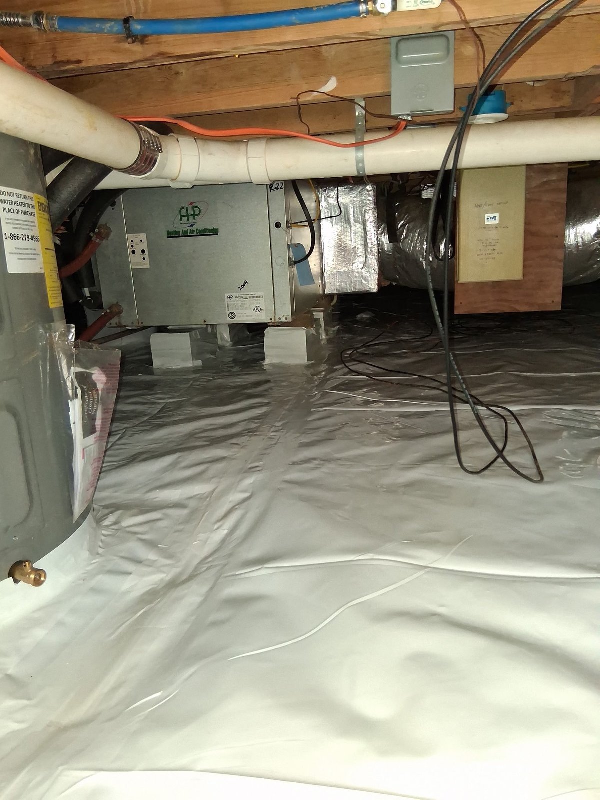 Crawl space under a house showing HVAC equipment, plastic vapor barrier, PVC pipes, and electrical wiring on wooden ceiling joists