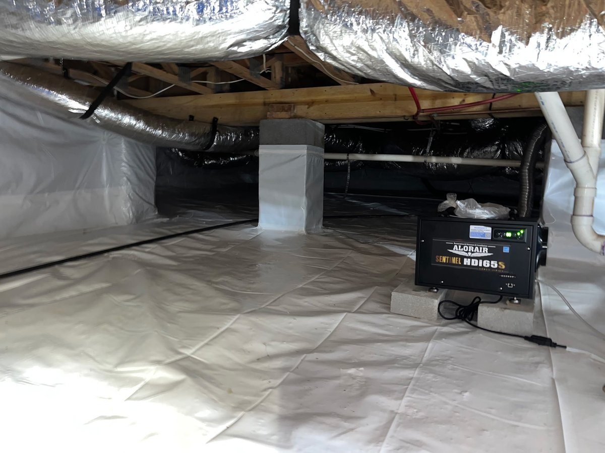 Crawl space interior with plastic sheeting, exposed wooden joists, black ductwork, and a dehumidifier unit on the floor