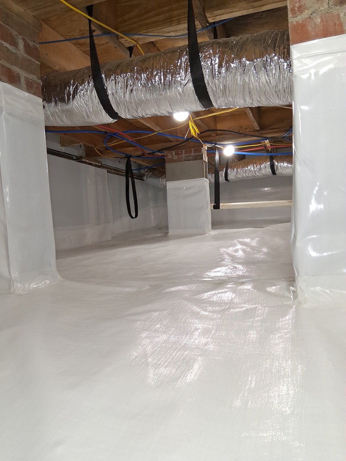 Unfinished basement with white plastic vapor barrier covering floor and walls, exposed wooden joists and ducts overhead