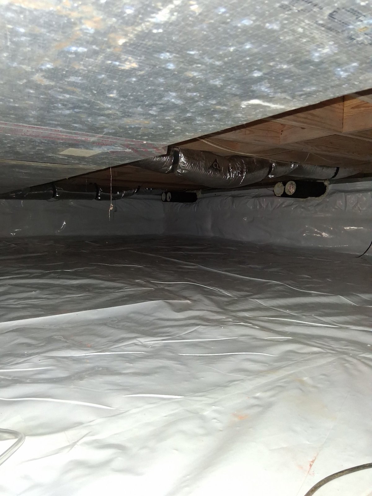 Underside of crawl space showing foundation supports, plastic sheeting, and exposed pipes